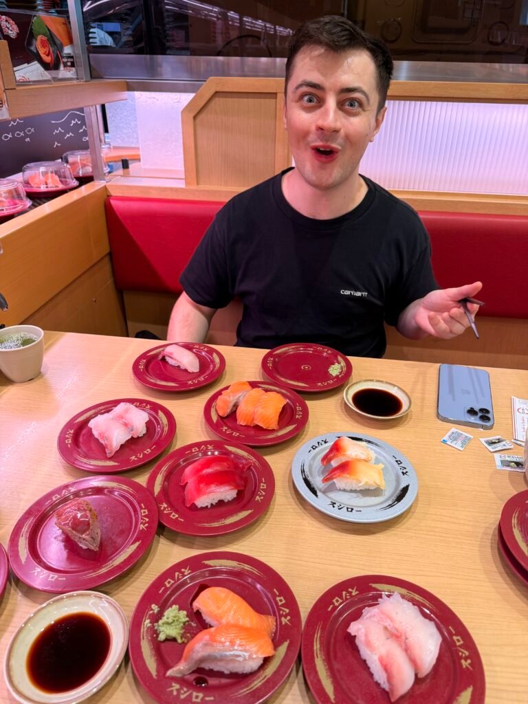 Man looking excited at a Bangkok kaiten conveyor belt sushi restaurant surrounded by multiple plates of salmon and tuna nigiri